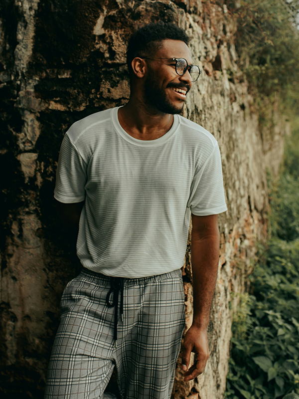 Man in his twenties in white shirt and jeans smiling while standing next to a wall outside